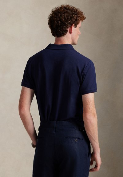 Navy polo shirt with a standard collar, short sleeves, and a relaxed fit, paired with dark trousers featuring rear pockets and buttons.