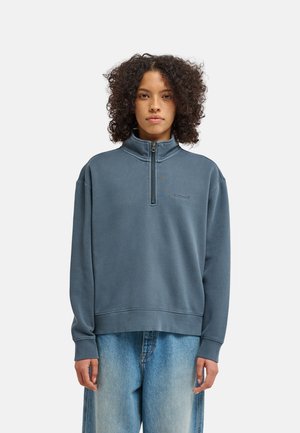 Femme aux cheveux bouclés portant un sweat à demi-fermeture éclair bleu et un jean ample bleu clair, debout devant un fond uni.
