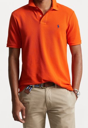 Orange polo shirt with a collar, short sleeves, and a blue logo on the chest. Paired with khaki pants and a multicolored striped belt.