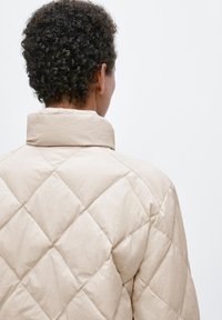 Beżowa pikowana kurtka z wysokim kołnierzem, z wzorem diamentowym i teksturowaną tkaniną, widziana od tyłu.