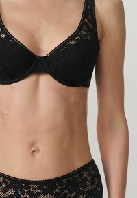 Femme portant un soutien-gorge en dentelle noire et une culotte en dentelle taille haute assortie, montrant son torse des épaules aux hanches sur un fond uni.