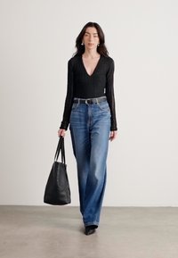 Musta ribbed pitkähihainen bodysuit, jossa on V-pääntie, yhdistettyinä löysiin sinisiin farkkuihin ja mustaan olkalaukkuun, kävelemässä neutraalilla taustalla.
