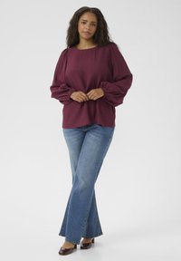 Burgunderfarbene Bluse mit langen Ärmeln und gerafften Schultern, kombiniert mit hellblauen, ausgestellt geschnittenen Jeans und dunkelbraunen Schuhen. Glattes, strukturiertes Material.