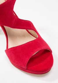 Sandale à talon haut en daim rouge à bout ouvert avec design découpé et semelle intérieure beige sur fond blanc.