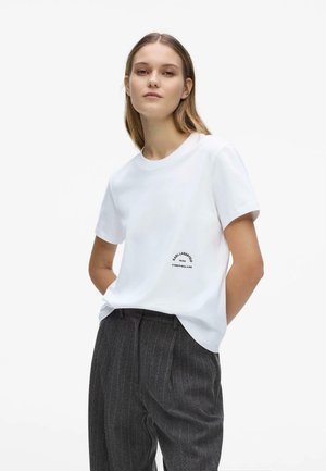 Jeune femme aux cheveux clairs portant une chemise blanche à manches courtes avec un petit texte et un pantalon gris à fines rayures, debout les mains derrière le dos.
