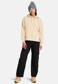 Beige hoodie med ficka framtill och dragsko, i kombination med svarta cargobyxor. Outfitten inkluderar en blårandig stickad mössa med tofs.