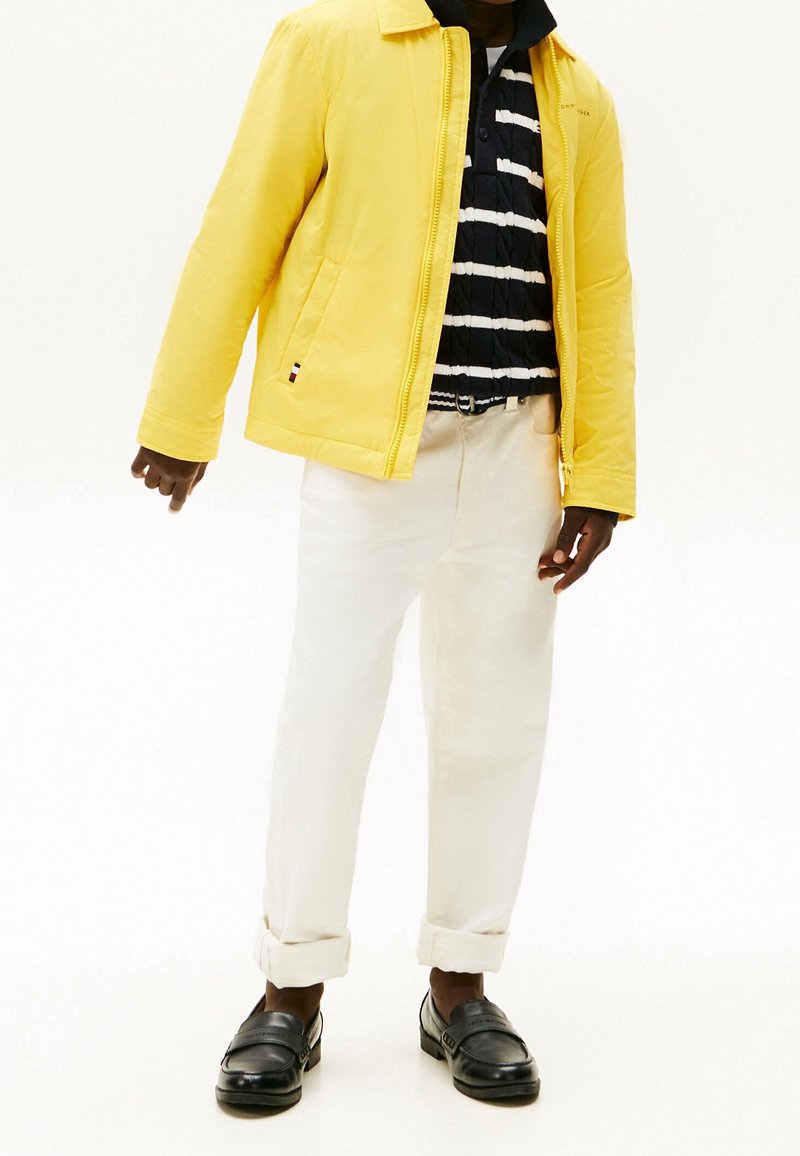 Personne vêtue d'une veste jaune, d'une chemise rayée noir et blanc, d'un pantalon blanc retroussé et de mocassins noirs, debout devant un fond uni.