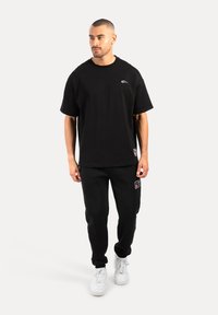 Zwarte katoenen T-shirt met ronde hals en korte mouwen, gecombineerd met zwarte joggingbroeken met zijzakken en een klein logo-patch. Witte sneakers.