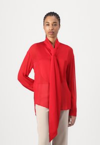 Rød chiffongbluse med lange ærmer og et knyttet skjerfdetalj ved halsen. Gjennomsiktig tekstur, løs passform og sideslits for bevegelse.