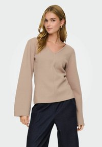 Femme aux longs cheveux blonds portant un pull beige à col V avec des manches larges et un pantalon bleu foncé, devant un fond uni.