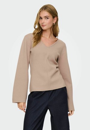 Mujer con cabello rubio largo vistiendo un suéter beige con escote en V y mangas amplias, y pantalones azul oscuro, frente a un fondo liso.