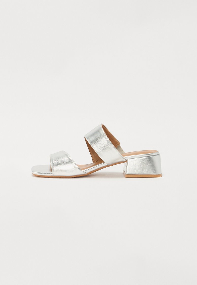 Stříbrné sandály na bloku s dvěma kříženými pásky. Hladký kovový povrch a jednoduchý design. Otevřená špička s nízkým čtvercovým podpatkem.
