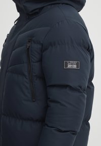 Marineblå dunjakke med glidelåslomme på siden, sømmerte paneler og logo-patch på overarmen. Glatt tekstur og isolert design.