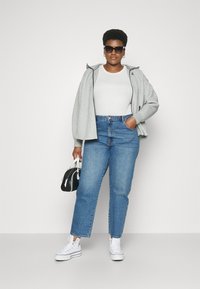 Sieviete ar saulesbrillēm, baltā kreklā, pelēkā kapuce, ziliem džinsiem, baltām augstpapēžu kedām, tur melnbaltu somu, stāvot pret vienkāršu fonu.