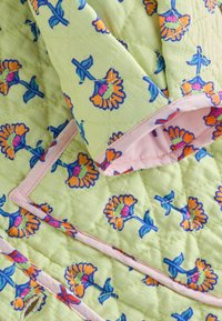 Tissu matelassé avec un fond vert clair orné de motifs floraux vibrants en orange et bleu. Bordé d'un galon rose doux.