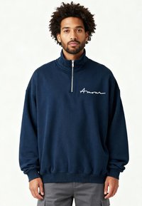 Sweat-shirt zippé bleu marine avec un col montant, manches longues, poignets et ourlet côtelés. Présente un texte cursif blanc « Amour » sur la poitrine.