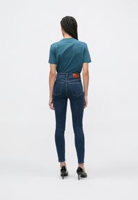 Modré úzké džínsy z denimu s vysokým pasem, s hnědou koženou nášivkou vzadu. Kombinovány s modrozeleným bavlněným tričkem a černými podpatky.