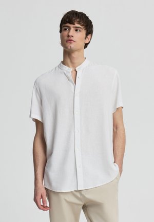 Giovane uomo che indossa una camicia bianca a maniche corte con collo alla coreana e pantaloni beige, in piedi con una mano in tasca.