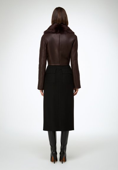 Giacca corta in pelle marrone con colletto in pelliccia, abbinata a una gonna nera di lana con due tasche posteriori. La modella indossa stivali neri alti.
