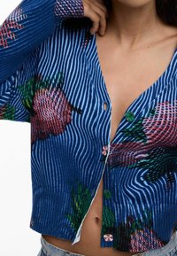 Gestrickter blauer Cardigan mit gerippter Textur, versehen mit einem floralen Muster in Rosa- und Grüntönen sowie kontrastierenden Knopfdetails.