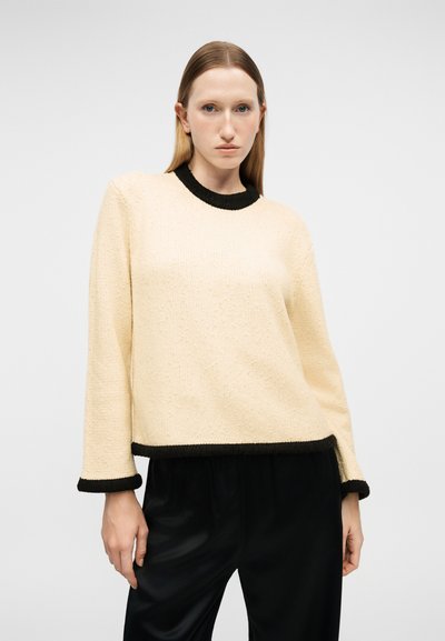 Femme aux cheveux raides portant un pull beige avec des bordures noires et un pantalon noir, debout devant un fond clair uni.