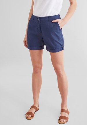 Femme portant un short bleu marine à ourlet roulé, un t-shirt blanc et des sandales marron, debout devant un fond uni gris clair.