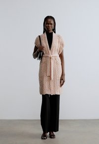 Απαλό ροζ πλεκτό ζακέτα με υφασμένο γεωμετρικό σχέδιο, με ζώνη στη μέση και κοντά μανίκια, σε συνδυασμό με μαύρο παντελόνι φαρδιάς γραμμής.