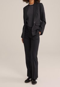 Černý tailored blazer přes černo-bílé pruhované tričko, doplněný černými kalhotami a černými teniskami s bílými pruhy.