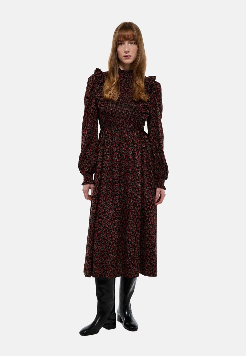 Robe noire avec motif floral, col haut, corsage smocké, manches longues et accents d'épaules froncés. Portée avec des bottes noires jusqu'aux genoux.