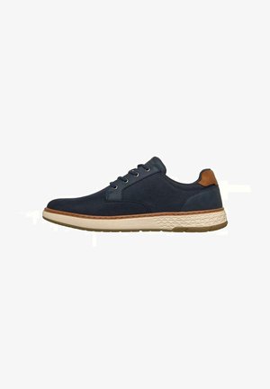 Zapato casual azul marino con cordones, lengüeta marrón en el talón, suela texturizada blanca y ribete beige, mostrado de lado sobre un fondo blanco.
