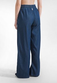 Osoba nosząca luźne, szerokie spodnie z ciemnoniebieskiego denimu z elastycznym pasem, stojąca na tle jednolitego białego tła.