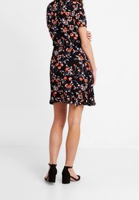 Robe florale noire avec des manches courtes, une taille ajustée et un ourlet évasé, rehaussée de fleurs orange et vertes. Portée avec des talons noirs.