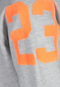 Tessuto grigio di felpa con una texture morbida, caratterizzato da grandi numeri arancioni "23" sulla manica in un materiale audace simile al feltro.