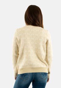 Suéter crema con un sutil patrón de logotipos texturizado, puños y dobladillo acanalados, combinado con jeans azules que presentan un diseño de bolsillo trasero curvado.