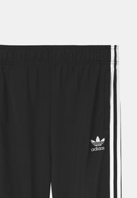 Černé Adidas tepláky s elastickým pasem, bílými bočními pruhy a vyšívaným logem Adidas Trefoil poblíž kapsy na bílém pozadí.