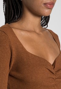 Top marrón con canalé y diseño de escote en V profundo, que presenta un detalle fruncido en el centro y una superficie texturizada.