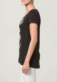 Woman wearing a black short-sleeve T-shirt with white graphic text, shown from the side, paired with white pants against a plain background.