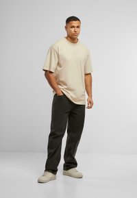 Beige cotton t-shirt with short sleeves, relaxed fit, and logo detail. Paired with black loose-fit jeans and beige sneakers. Plain background.