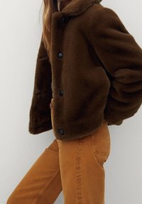 Brązowa kurtka z krótkiego sztucznego futra z kołnierzem, posiadająca czarne guziki oraz puszystą teksturę. Połączona z pomarańczowymi dżinsami.