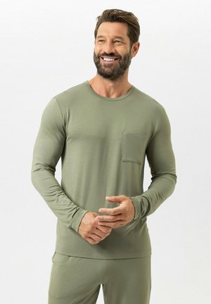 Mann mit Bart lächelnd, trägt olivgrünes langärmeliges Hemd mit Brusttasche und passende Hose, steht vor einem einfarbigen weißen Hintergrund.