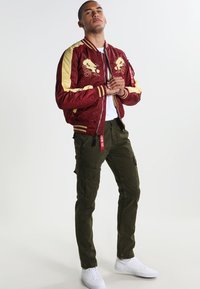Chaqueta bomber roja con detalles y bordados en dorado, combinada con pantalones cargo oliva y zapatillas blancas. Tejido suave, corte atlético y cremalleras.