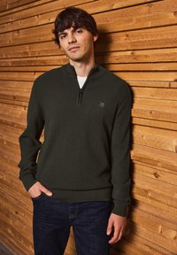 Un sweat zippé vert foncé avec un motif côtelé texturé, porté sur une chemise blanche, associé à un jean bleu foncé, le tout devant un fond en bois.