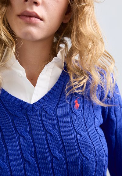 Zils adītais džemperis ar V-veida kakla izgriezumu un mazu sarkanu logotipu. Valkāts virs baltas krekliņa ar apkakli, izceļot teksturētu audumu.