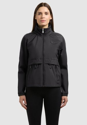 Kobieta nosi czarną kurtkę zapinaną na zamek z wysokim kołnierzem, przednimi kieszeniami i ściągaczem w talii, na czarnych spodniach, na tle jednolitego jasnego tła.