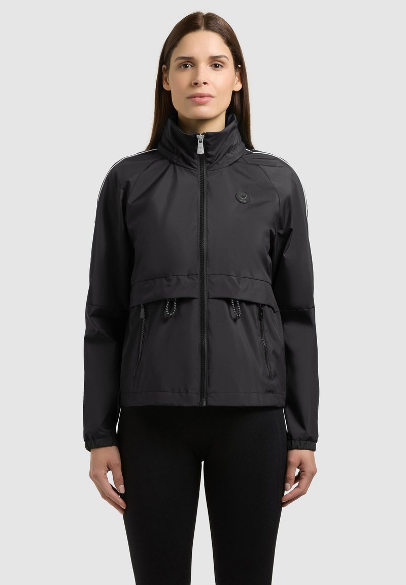 Femme portant une veste noire zippée avec col montant, poches avant et taille avec cordon de serrage, associée à un pantalon noir, sur un fond clair uni.
