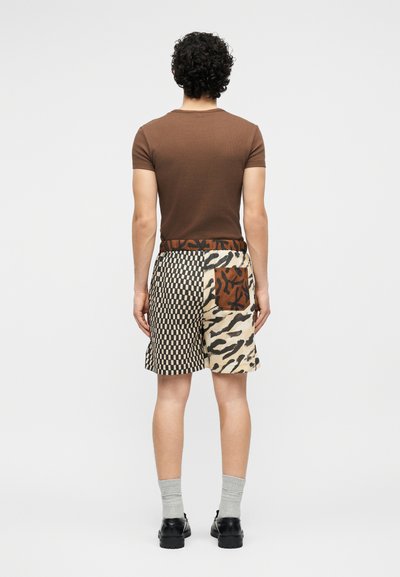 Homme aux cheveux bouclés portant un t-shirt marron et un short patchwork avec des motifs à carreaux et des motifs abstraits d'animaux, debout dos à la caméra.