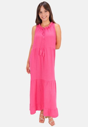 Junge Frau lächelt, trägt ein ärmelloses, knallpinkes Maxikleid mit Quastenbändern und weiße Riemchensandalen auf einfarbigem Hintergrund.