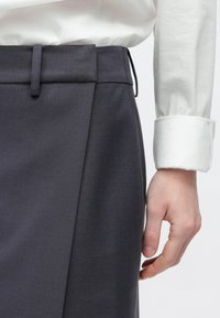 Roka atpūšas pie tumši pelēkas pielāgotas svārka ar jostas cilpām, kopā ar baltu garām piedurknēm krekls ar uzlocītu aproci.