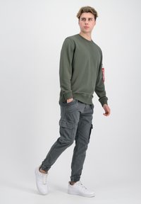 Sudadera verde con cuello redondo y detalles de etiqueta roja, combinada con pantalones cargo grises con bolsillos y puños ajustados, usados con zapatillas blancas.
