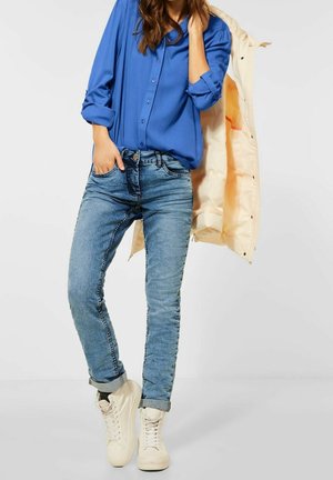 Kobieta w niebieskiej koszuli na guziki, jasnych podwiniętych dżinsach, kremowych trampkach, trzymająca beżowy płaszcz przewieszony przez ramię na białym tle.